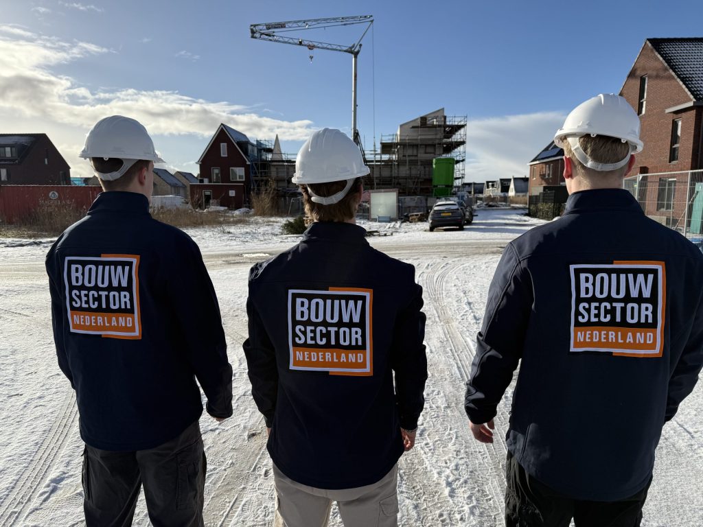 Drie vakmensen in werkjassen met logo van Bouwsector Nederland kijken naar rijen nieuwe woningen